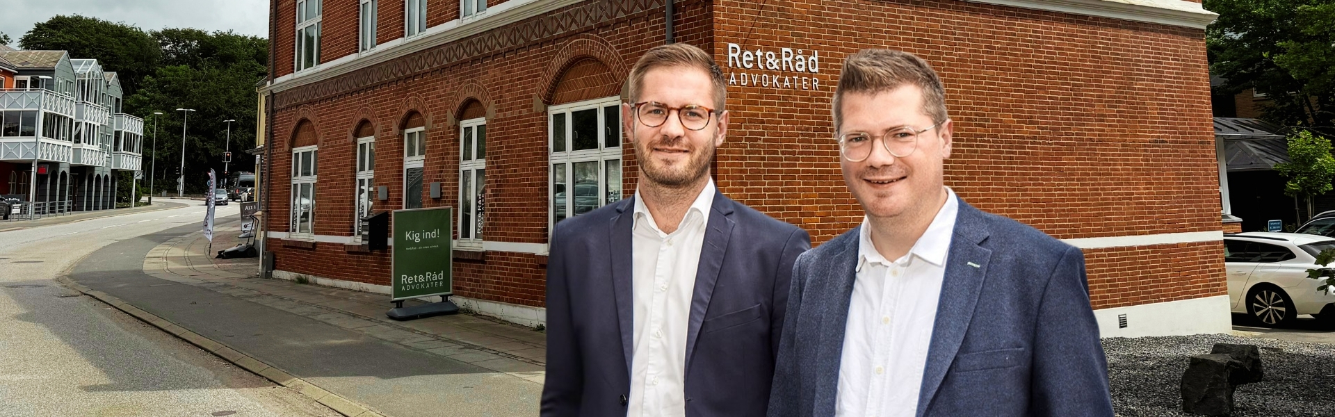 To mænd iført jakkesæt står foran en rød murstensbygning med skiltet "Ret&Råd ADVOKATER" på væggen og et grønt skilt ved fortovet med teksten "Kig ind!".