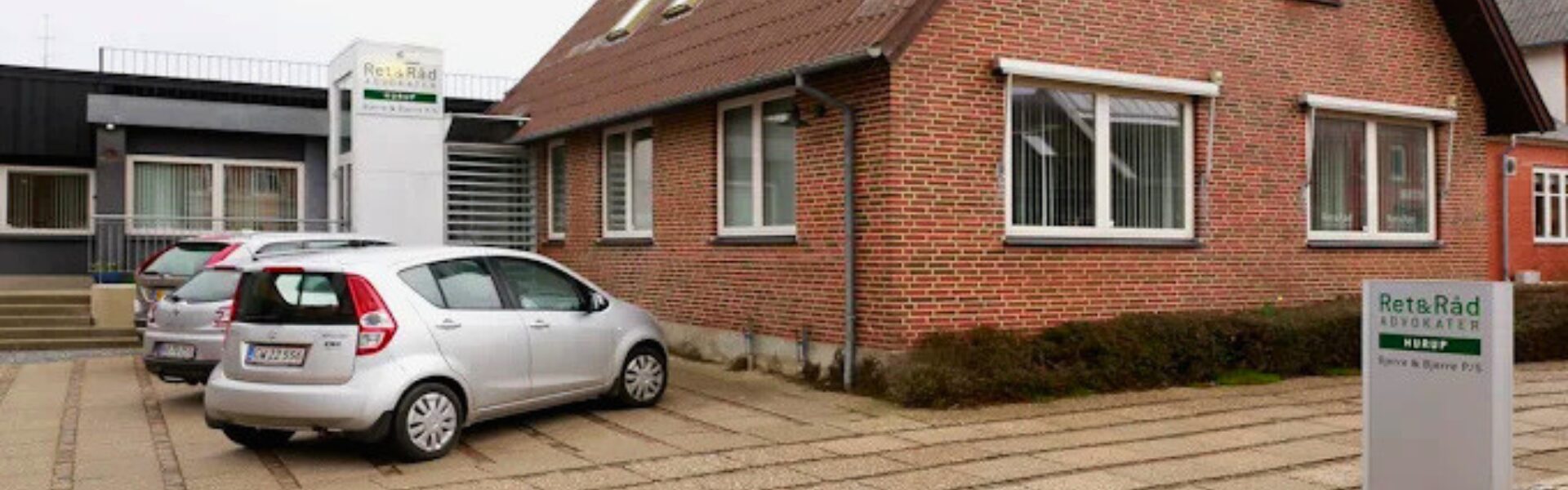 Sølvfarvet bil parkeret på en flisebelagt indkørsel foran en rød murstensvilla med tre hvide rammevinduer, et lille bed med lave buske og et nedløbsrør under et rødt tegltag.