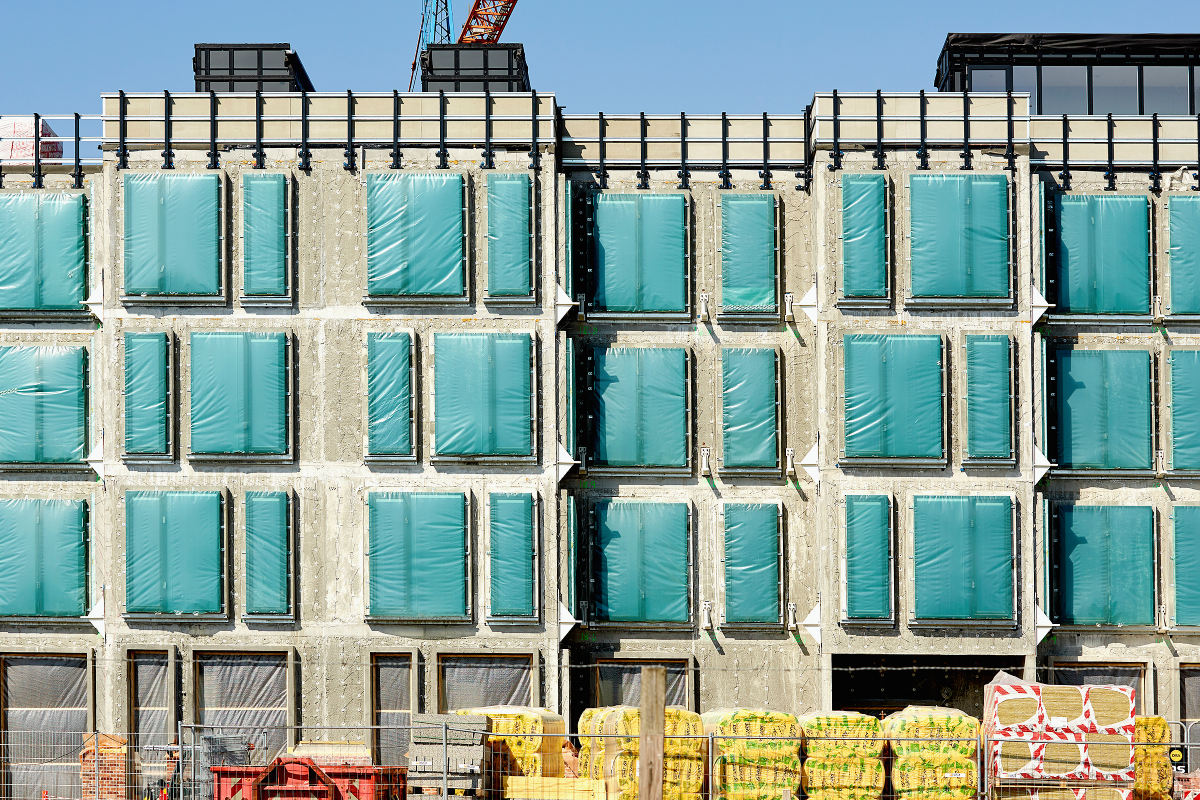 Bygning under konstruktion med vinduer dækket af grøn plastfolie. Foran bygningen ses byggepladsudstyr og stakke af isoleringsmateriale.