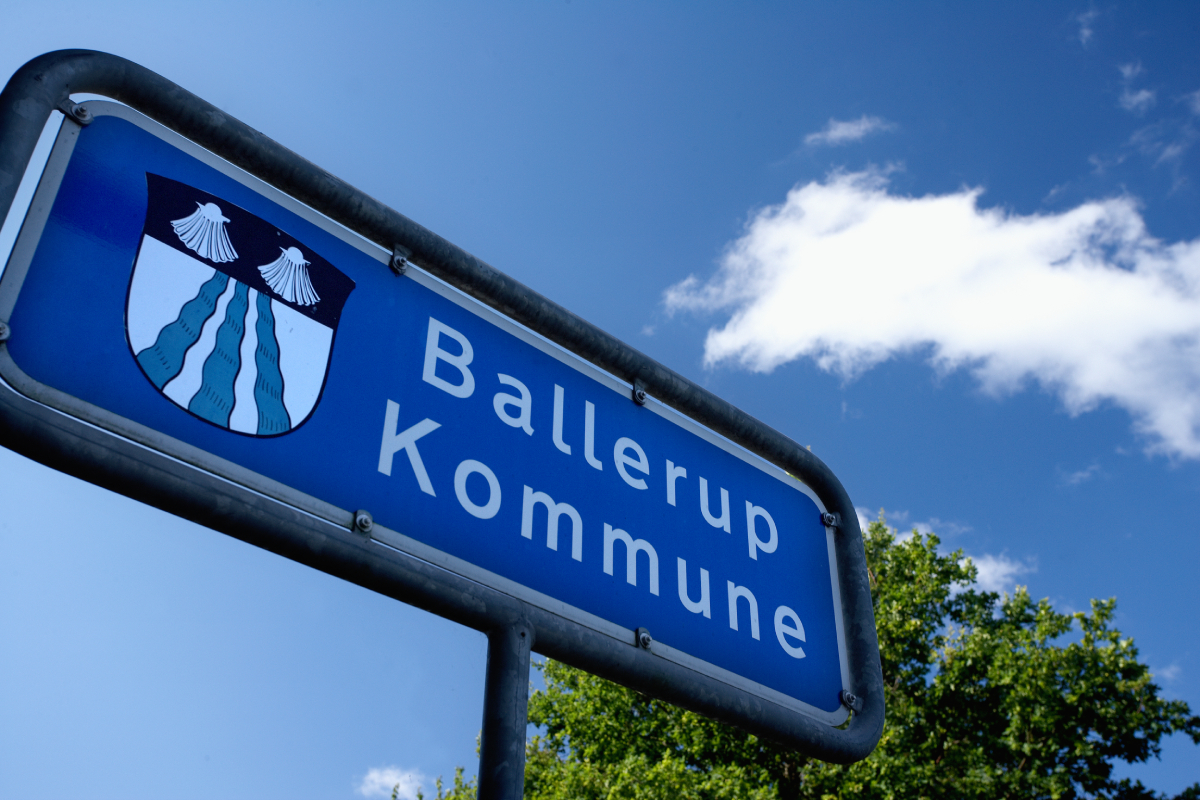 Et blåt skilt med teksten "Ballerup Kommune" og et våbenskjold med to muslingeskaller og bølgende linjer, set mod en blå himmel med hvide skyer.