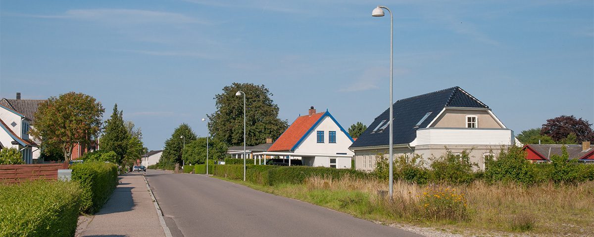En landsbyvej med huse, herunder et med rødt tag og en hæk foran.