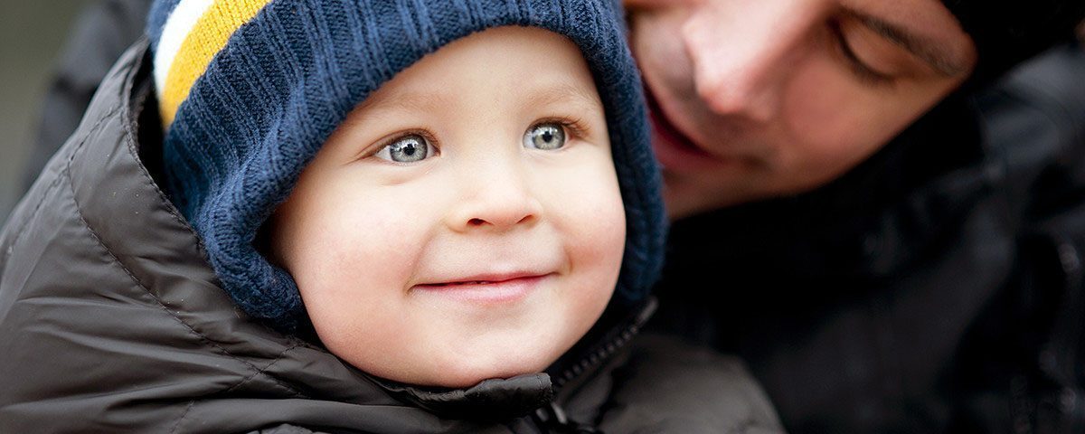 Et lille barn med blå hue smiler, mens en voksen er delvist synlig ved siden af.