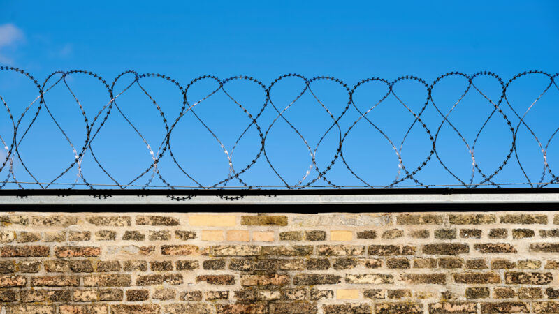 Murstensmur toppet med pigtråd, under en klar blå himmel.