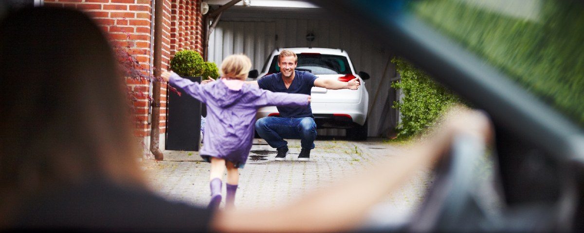 Et barn løber hen mod en voksen, der knæler med åbne arme foran en bil i en garage.