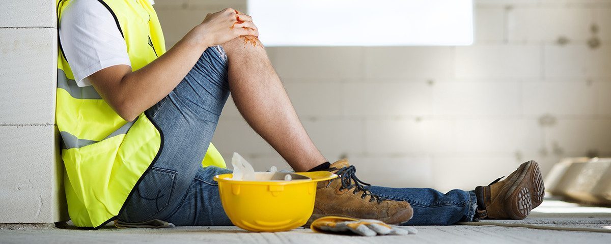 3. Person sidder med krydsede ben ved siden af en gul sikkerhedshjelm og arbejdshandsker på en byggeplads.