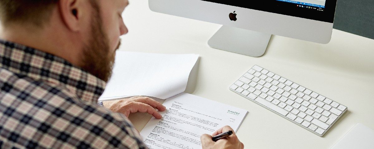 En person sidder ved et skrivebord med en computer og udfylder papirer med en pen.
