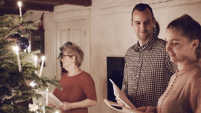 To personer holder papirer og står ved siden af et juletræ i en hyggelig stue.