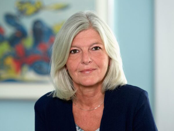 En person med skulderlangt, hvidt hår iført en mørkeblå jakke. Baggrunden er en væg med et farverigt abstrakt maleri.