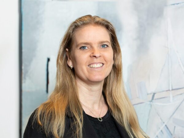En person med langt blondt hår iført en sort bluse og halskæde står foran en abstrakt malet baggrund i lyse nuancer.
