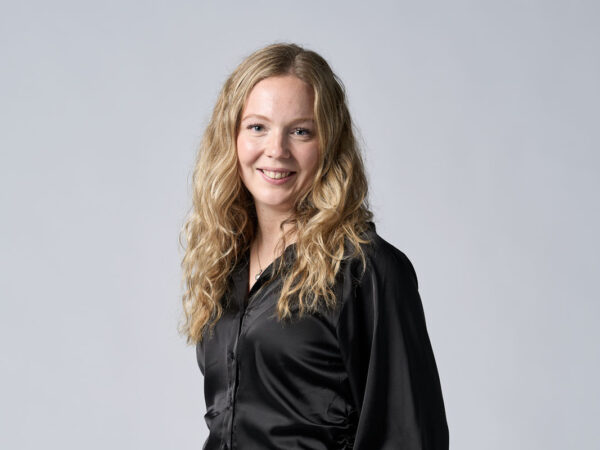 En kvinde med langt, bølget blond hår iført en sort skjorte smiler foran en ensfarvet, grå baggrund.