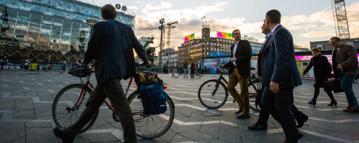 Folk cykler og går på en plads med bygninger oplyst af farvet lys i baggrunden.