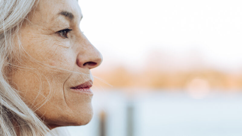 Langt, bølget blond hår set fra siden med en sløret baggrund af vand og træer.