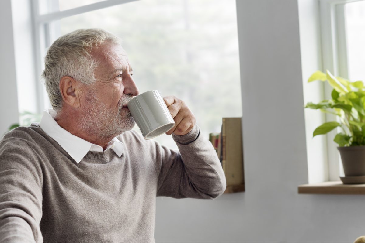 En ældre mand sidder ved et vindue og drikker kaffe fra en hvid kop.