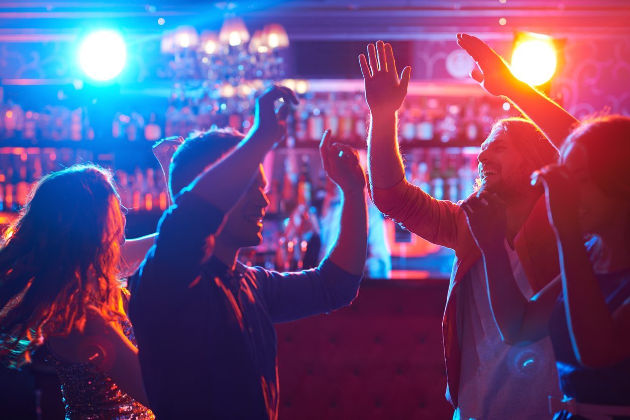 Folk danser under farverige lys på en natklub med en bar i baggrunden.
