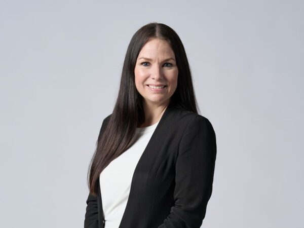 En person med langt, mørkt hår iført en sort blazer og hvid bluse står foran en ensfarvet, lys grå baggrund. Ansigtet er sløret.