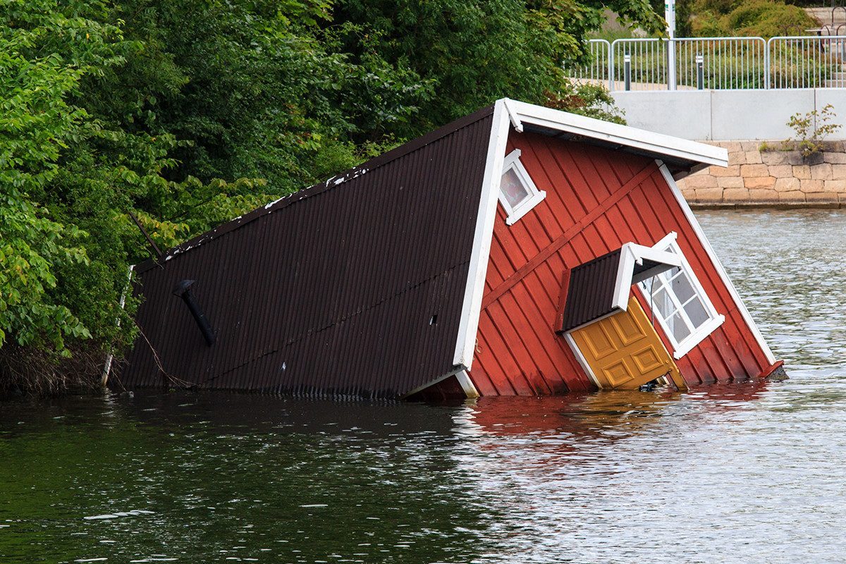 Et rødt hus ligger delvist under vand, omgivet af træer og en vej i baggrunden.