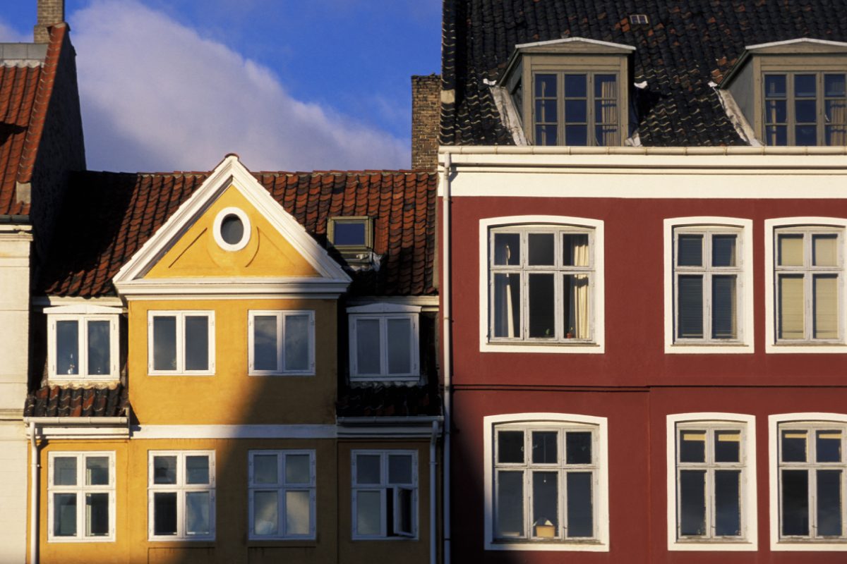To farverige bygninger, en gul og en rød, med klassiske vinduer står side om side under sollys.