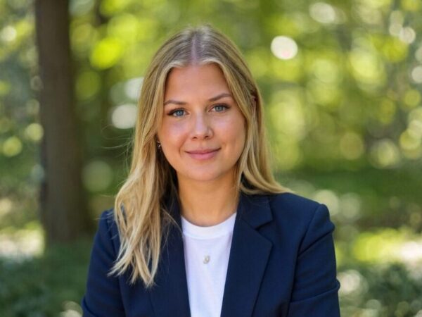 En kvinde med blondt hår er iført en marineblå blazer og hvid T-shirt. Baggrunden viser en skov med grønne træer og sollys.