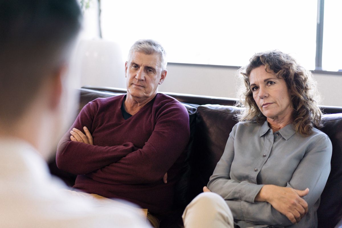 To voksne personer sidder på en sofa; den ene har krydsede arme, og den anden ser ned.