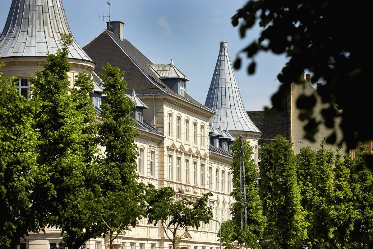 En bygning med europæisk arkitektur, omgivet af grønne træer under en klar himmel.