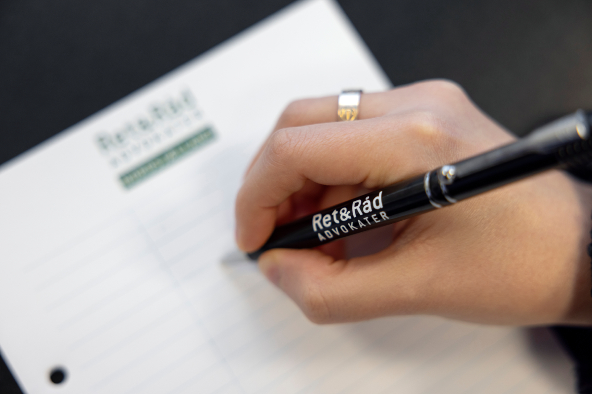 En hånd med en ring holder en sort kuglepen med teksten "Ret&Råd ADVOKATER" og skriver på en linjeret notesblok med logoet "Ret&Råd ADVOKATER" i øverste venstre hjørne.