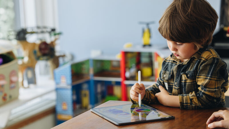Et barn iført en ternet skjorte tegner med en stylus på en tablet, mens en voksen hånd ses ved siden af. Baggrunden viser legetøj og en legehusreol i et lyst rum.