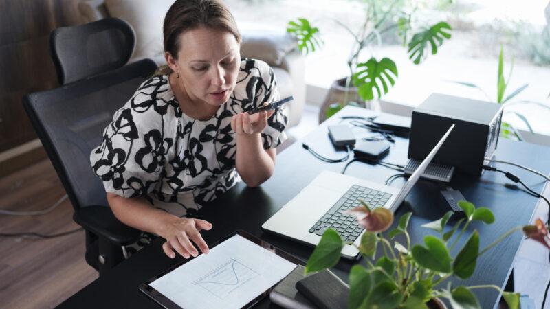 En kvinde sidder ved et skrivebord og taler i en smartphone. På bordet ligger en tablet med en graf, en bærbar computer og planter i forgrunden.