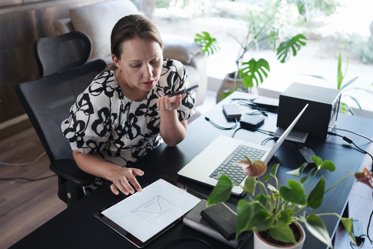 En kvinde sidder ved et skrivebord og taler i en smartphone. På bordet ligger en tablet med en graf, en bærbar computer og planter i forgrunden.