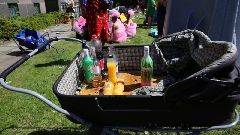 En barnevogn fyldt med flasker, herunder juice og alkohol, på en græsplæne under en udendørs begivenhed med mennesker iført farverige kostumer i baggrunden.