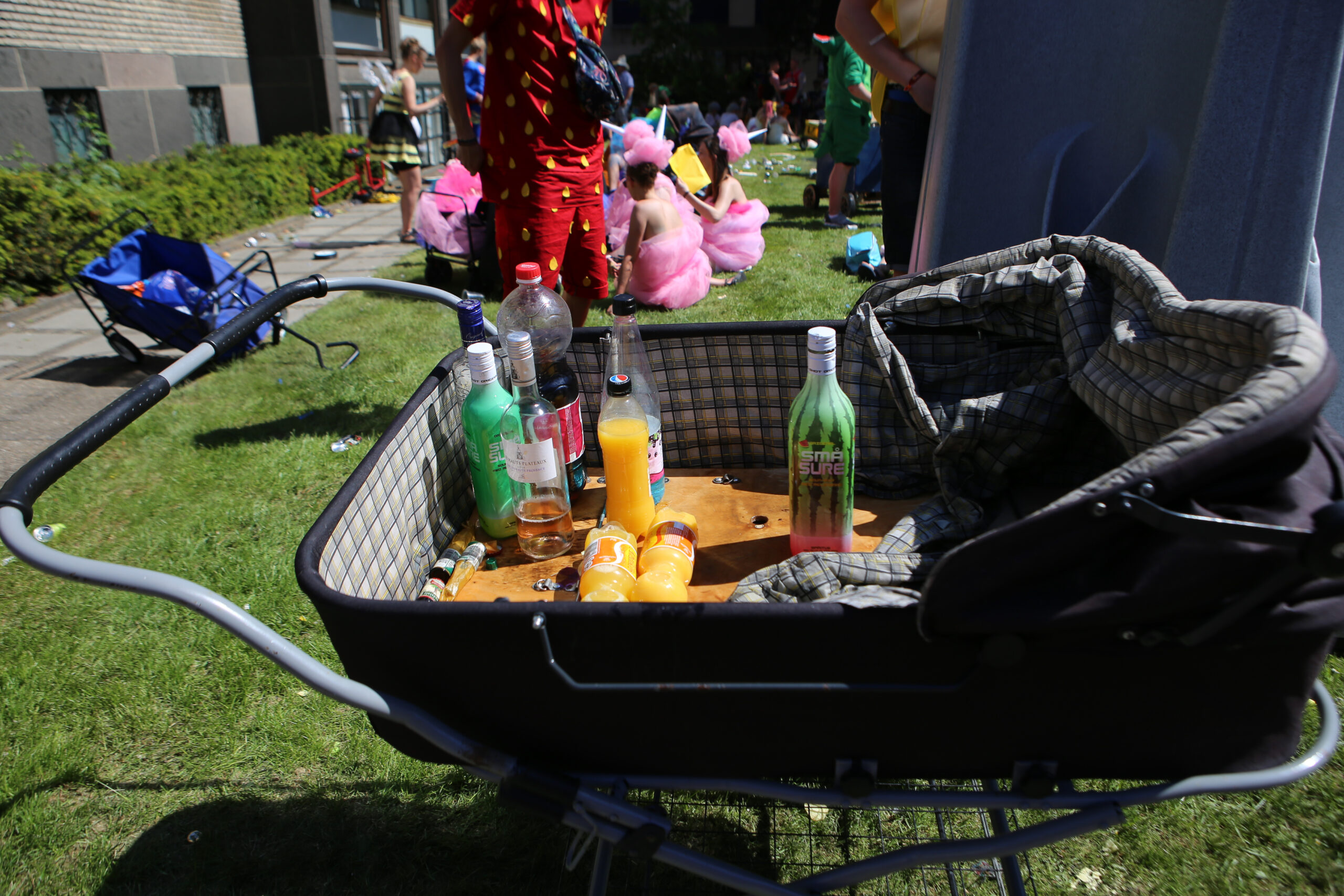 En barnevogn fyldt med flasker, herunder juice og alkohol, på en græsplæne under en udendørs begivenhed med mennesker iført farverige kostumer i baggrunden.
