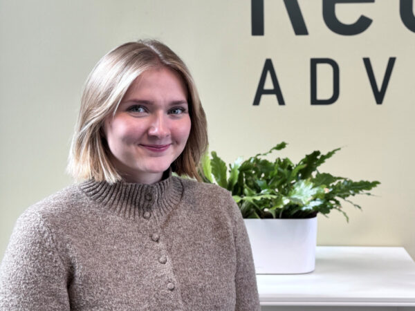 En kvinde med kort, lyst hår smiler foran en lys væg med teksten "Ret ADVO." En grøn plante i en hvid potte står på en hvid overflade i baggrunden.