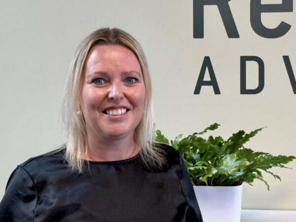 En kvinde med lyst hår og sort bluse smiler foran en væg med teksten "ADV" og en plante i en hvid krukke.