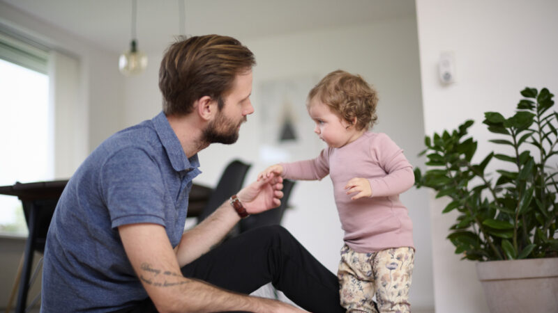 En voksen person i en blå polo trøje sidder på gulvet og holder hånden med et lille barn i lyserød bluse og mønstrede bukser. En stor grøn plante ses i baggrunden.